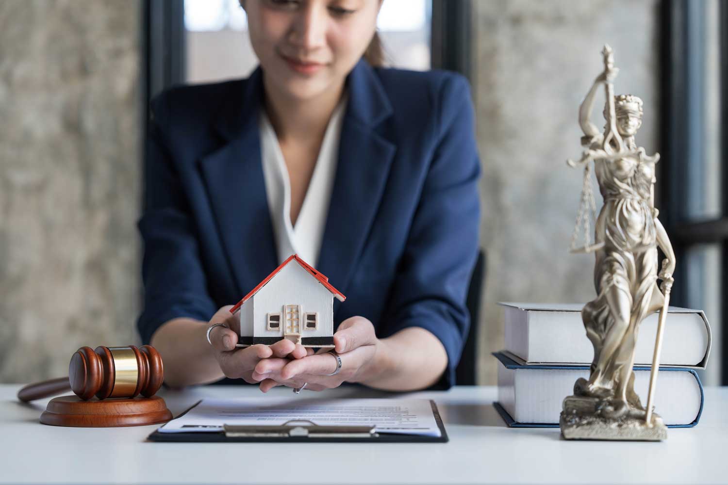 A beutiful girl Law Firm holding a house model, symbolizing property law, with legal books and a statue of justice in background.